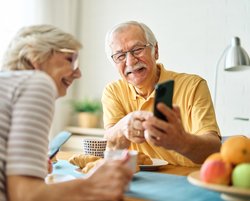 Zwei ältere Menschen sitzen an einem gedeckten Tisch. Der ältere Herr hat ein Smartphone in der Hand und zeigt der Frau etwas darauf. Beide schauen fröhlich.