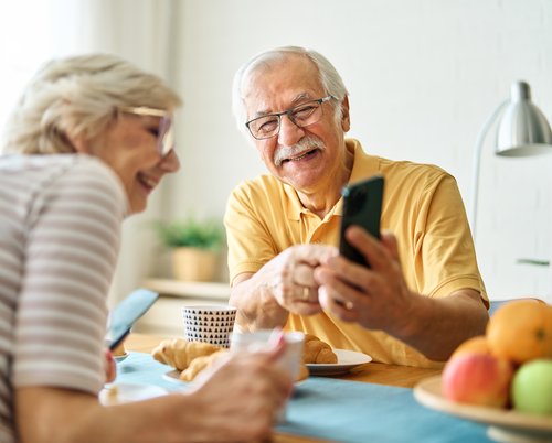 Zwei ältere Menschen sitzen an einem gedeckten Tisch. Der ältere Herr hat ein Smartphone in der Hand und zeigt der Frau etwas darauf. Beide schauen fröhlich.