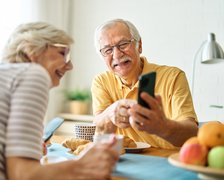 Zwei ältere Menschen sitzen an einem gedeckten Tisch. Der ältere Herr hat ein Smartphone in der Hand und zeigt der Frau etwas darauf. Beide schauen fröhlich.