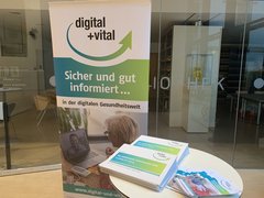 Ein Aufsteller des Projektes vor einer Glaswand. Darauf sind das Projekt-Logo und der Slogan "Sicher und gut informiert in der digitalen Gesundheitswelt". Davor ein Stehtisch mit Materialien des Projektes.